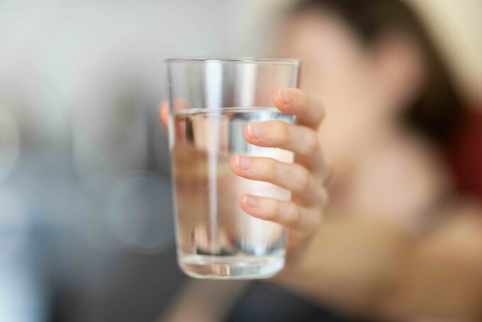 Eine Hand hält ein Glas mit klarem Wasser. Der Hintergrund ist unscharf, wodurch der Fokus auf dem Glas liegt.